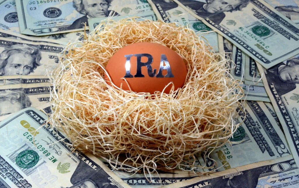 An egg that has the letters IRA on it to symbolize Individual retirement Account in a as a nest egg on top of twenty dollar bills spread out beneath it.
