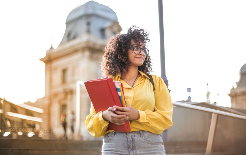 A young adult woman college student standing in front of the school holding her college books.