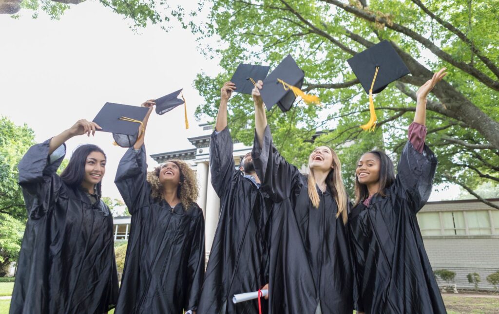 A photo of college graduate young adults wearing graduation gowns and tossing their caps in the air.