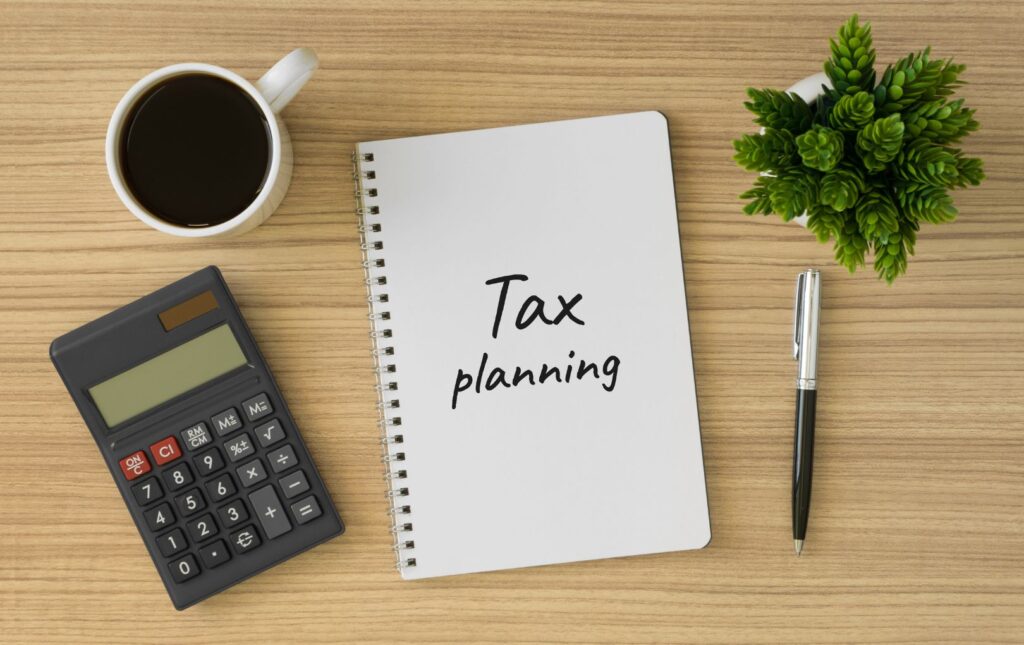 "Tax Planning" notebook on a table with a cup of coffee, calculator, pen and green plant on a desk.