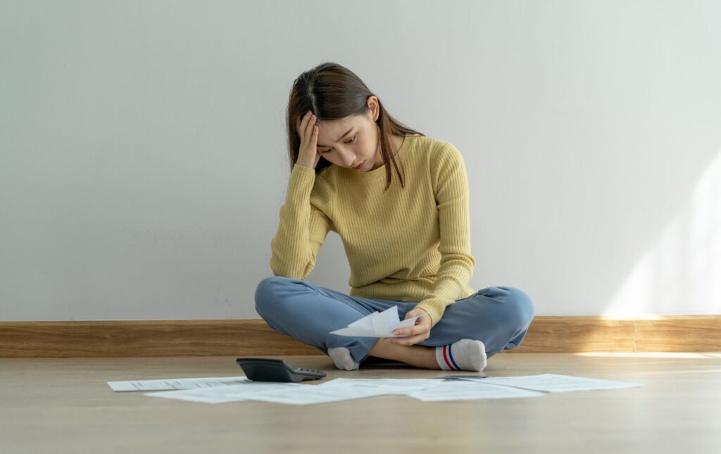 young woman sitting on the floor in her apartment looking down at her bills as she is living paycheck-to-paycheck